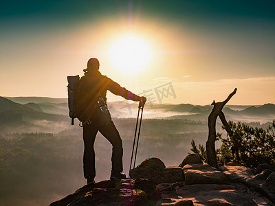 背着背包的徒步旅行者靠在登山杖上凝视着朦胧的日出。