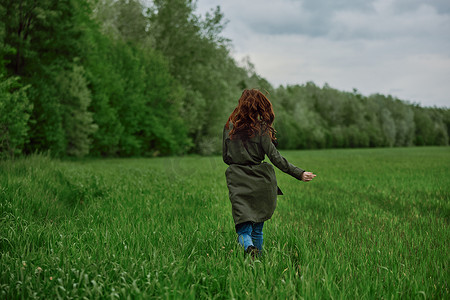 春天多云的天气里，一位身穿长雨衣的女士在高高的绿草丛中跑过田野