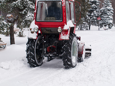 除雪摄影照片_拖拉机在降雪后从城市公园的人行道上除雪