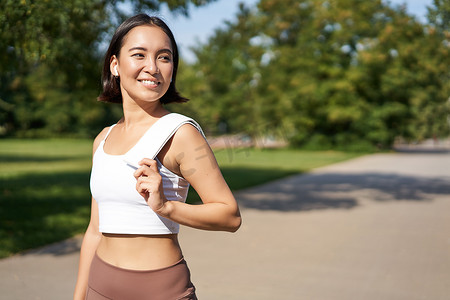 小儿出汗摄影照片_微笑的亚洲健身女孩肩上拿着毛巾，在公园锻炼，在户外训练后出汗