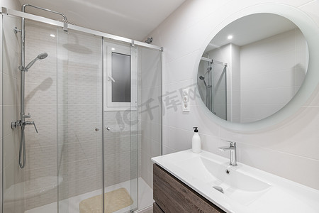 白色窗户摄影照片_白色瓷砖浴室，墙上有大圆镜，反射带玻璃栏杆和金属配件的淋浴区。