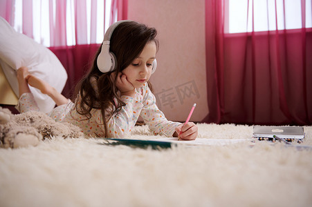 戴着无线耳机和睡衣的可爱小女孩躺在卧室的地毯上，在她的相册里画画