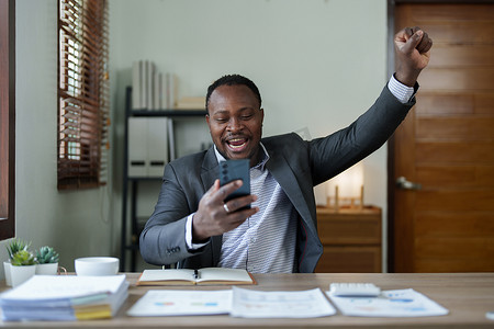 中年男子美国非洲商人手持智能手机，双手举起赢家，这是一种姿态，很高兴成功庆祝成就成功