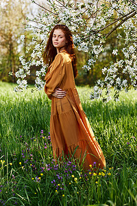 一个苗条、甜美的女人穿着橙色长裙站在一棵开花的树旁高高的草丛中，双臂交叉放在胸前，望向远方