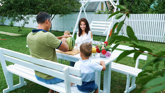 家庭餐摄影照片_夏天，一个年轻的家庭和两个有趣的孩子坐在花园的餐桌旁。