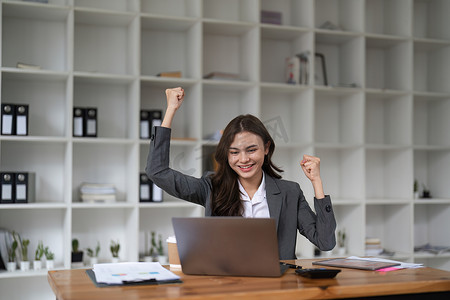 快乐的女上班族感到兴奋，举起拳头庆祝职业阶梯晋升或奖励，坐在办公桌前的女商人接收在线新闻，取得成功的成功工作理念