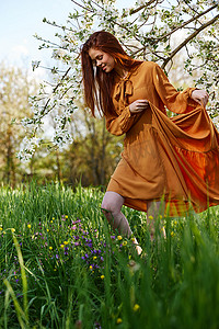 绿色树苗条摄影照片_一个快乐、苗条、甜美的女人穿着橙色长裙站在一棵开花的树旁高高的草丛中，开心地微笑着掀起裙子的下摆