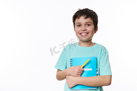黄色青春摄影照片_聪明的小学生，聪明的男孩，拿着课本和黄色笔，对着镜头微笑，白色背景