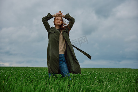 一位身穿长大衣的女士站在多云的田野里，背对着风，双手举在脑后