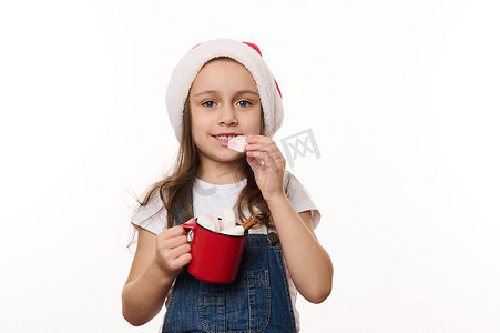 可爱的小女孩的白色孤立肖像，戴着圣诞帽，拿着红杯热巧克力，吃着棉花糖
