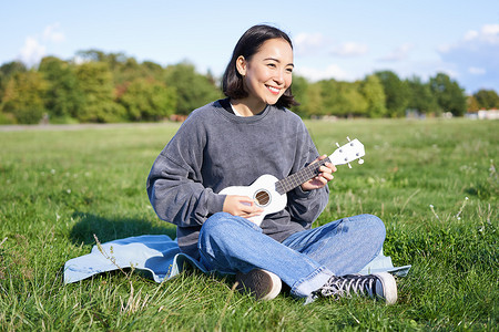 阳光明媚的春日，美丽的亚洲女孩坐在公园里弹尤克里里唱歌，在户外放松