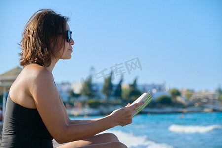 坐在海边沙滩上的太阳镜泳装阅读书的成熟女性