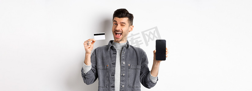 开朗的帅哥展示优惠，空荡荡的智能手机屏幕和塑料信用卡，向你眨眼微笑，推荐报价，白色背景