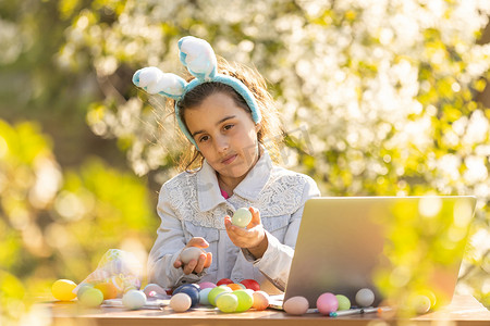 快乐的少女戴着兔耳朵在花园里用笔记本电脑画复活节彩蛋