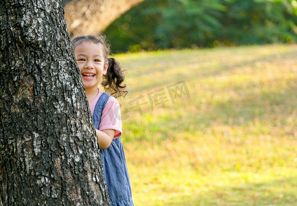 亚洲小女孩的画像躲在树后，在公园或花园里微笑，玩躲猫猫。
