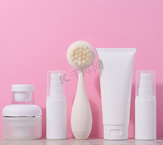 白色塑料管和奶油罐，以及粉红色背景中用于面部清洁的按摩刷，用于美容程序的物品
