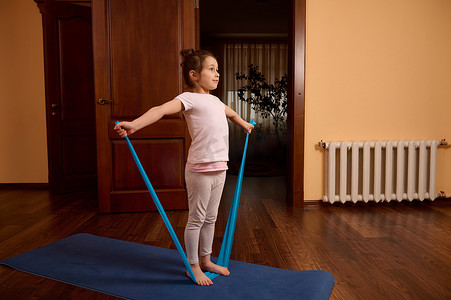 运动健身垫摄影照片_穿着运动服的小女孩站在健身垫上，在家里用松紧带做手臂伸展运动