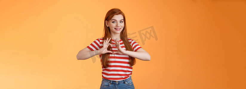 可爱可爱开朗的红发女友表达爱和珍惜关系，庆祝周年纪念展示心形标志，温柔地微笑，表白同情，站在橙色背景