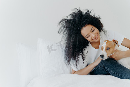 可爱照片墙摄影照片_可爱的非裔美国女孩在与狗一起醒来后躺在床上休息的室内照片，与宠物共度时光，坐在舒适的床上靠着白墙。