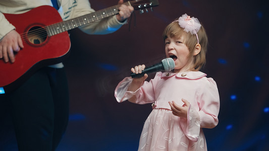 穿着复古裙子的小女孩在舞台上唱歌，她的父亲弹着原声吉他