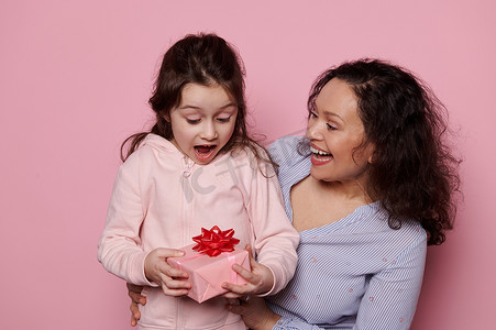 快乐的小女孩在从她慈爱的母亲那里得到生日礼物时表达惊喜，她被隔离在粉红色背景中