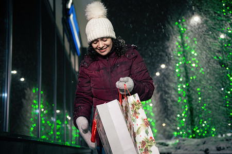 迷人的女人提着购物袋，走在街上，在冬天下雪的夜晚，节日的灯光照亮了她。