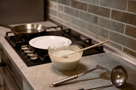 玻璃碗中的木勺，配煎饼面糊，厨房台面上的不锈钢厨房用具，黑炉上的白盘和煎锅