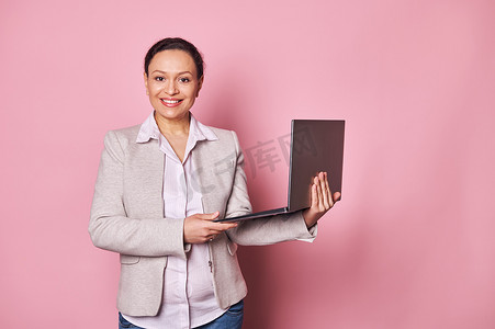 成熟的女商人微笑着，自信地看着相机，拿着笔记本电脑，背景是粉红色的。