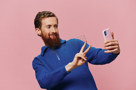 粉色主背景摄影照片_一位手持手机的红头发男子的肖像，他在手机上自拍和拍照，在粉红色背景下微笑，博主