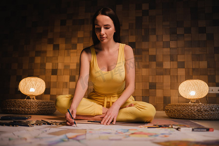 纸荷花摄影照片_一个穿着黄色衣服的女孩坐在莲花位置上，在家里用晚上的灯光在纸上画画