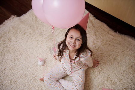 顶视图可爱的小孩，穿着睡衣和粉色派对帽的生日女孩，坐在地毯上，微笑着看着镜头
