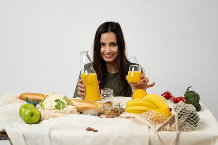 身穿绿色 T 恤的女人将玻璃瓶中的果汁倒在桌子上，桌子上放着网眼生态袋、健康的素食蔬菜、水果、面包、零食。