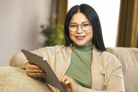 自信的亚洲中年女性享受在家放松的时刻，在平板电脑上轻松舒适地浏览互联网