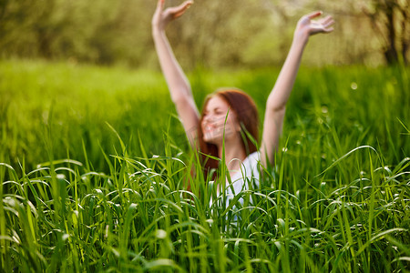 快乐、心满意足的女人坐在高高的绿草上，高高举起双臂享受愉快的一天