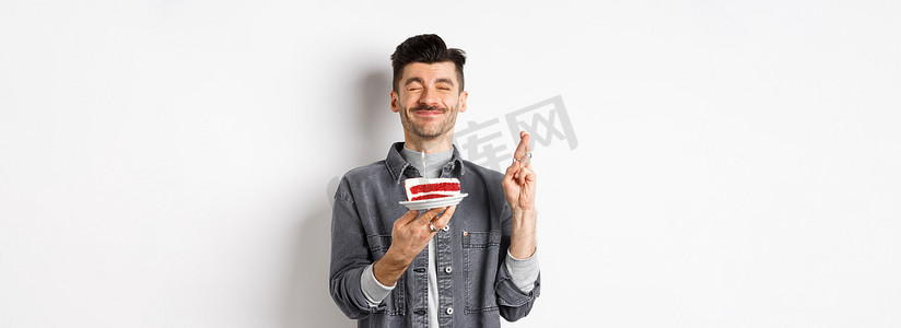 满怀希望的生日小伙子用手指交叉许愿，在派对上拿着生日蛋糕，站在白色背景下