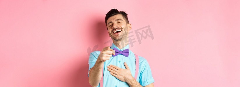 戴领结的帅哥用手指指着镜头，嘲笑一些有趣的事情，查看搞笑的宣传片，站在粉红色背景中