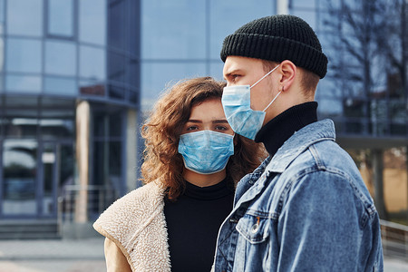 一对戴着防护面具的夫妇在检疫时间在商业大楼附近的城市户外散步。