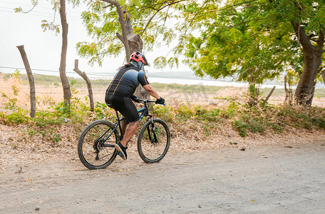 胖运动摄影照片_在被树木包围的土路上胖骑自行车的人，在路上胖乎乎的骑自行车的人的生活方式，在土路上的专业骑自行车的人
