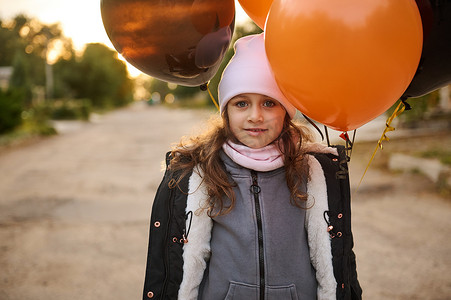 漂亮的小女孩带着黑色和橙色的气球走在街上。
