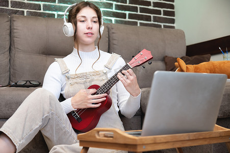 年轻漂亮的女人正坐在家里的地板上，学习弹吉他或四弦琴，站在笔记本电脑和一杯咖啡旁边。