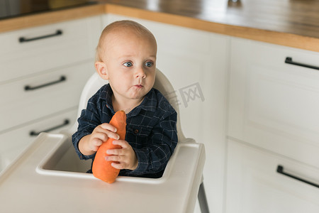 婴儿喂养摄影照片_坐在婴儿椅上的男婴在厨房背景复制空间吃胡萝卜 — 婴儿喂养概念