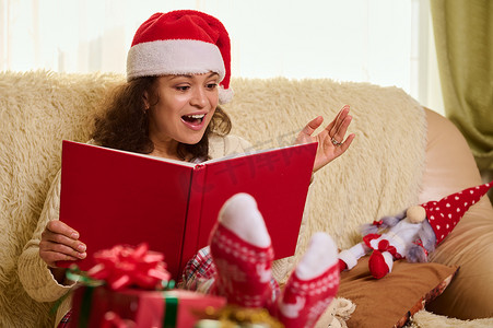 寒假阅读摄影照片_美丽的西班牙裔女人戴着圣诞老人的帽子和圣诞图案的袜子，在寒假里读童话故事。