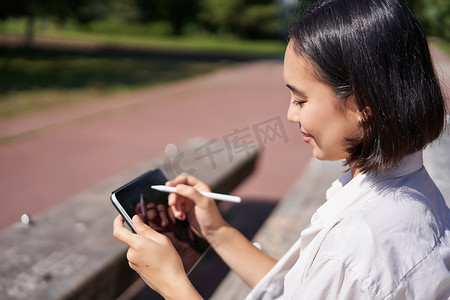 亚洲年轻女性在公园里呼吸新鲜空气的画像，坐在平板电脑和数字笔旁，开心地笑着