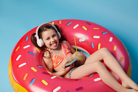 儿童游泳圈摄影照片_戴着无线耳机调皮的小女孩，在手机上玩得开心，躺在充气粉色甜甜圈游泳圈上