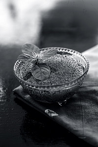 将浸泡过的 sabja 种子或 falooda 种子或甜罗勒种子放在木质表面棕色餐巾纸上的玻璃碗中，里面有一些薄荷叶。用于许多调味饮料。