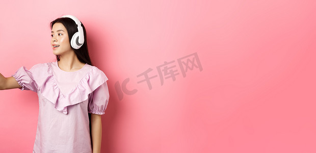 美丽的亚洲女博主戴着无线耳机自拍，在智能手机上为社交媒体拍照，穿着粉色背景的裙子