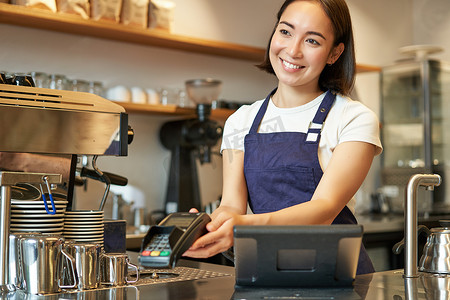 微笑的亚洲女咖啡师，给客户 POS 终端，帮助用信用卡支付，站在咖啡店柜台，在咖啡馆工作