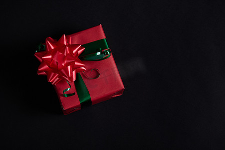 圣诞礼物放在礼盒里，礼盒里裹着红色包装纸，黑色背景上有绿色丝带和红色系结蝴蝶结
