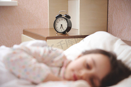 床上摄影照片_专注于显示六点半的黑色闹钟，在床边床头柜上，在一个熟睡的女童的模糊前景中