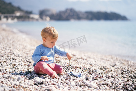 穿着蓝色衬衫和红色裤子的小孩坐在卵石滩上，手里拿着一根棍子